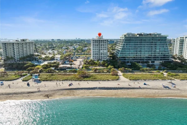 Aerial view of a beach with turquoise water and a row of modern buildings; a red location pin sits on top of a white mid-rise.