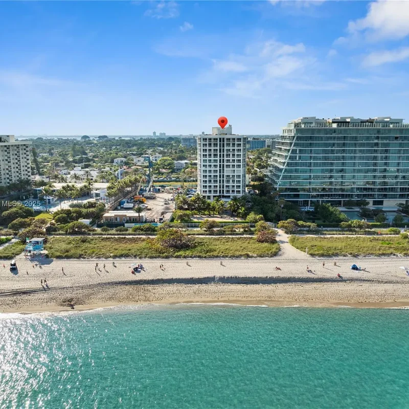 Aerial view of a beach with turquoise water and a row of modern buildings; a red location pin sits on top of a white mid-rise.