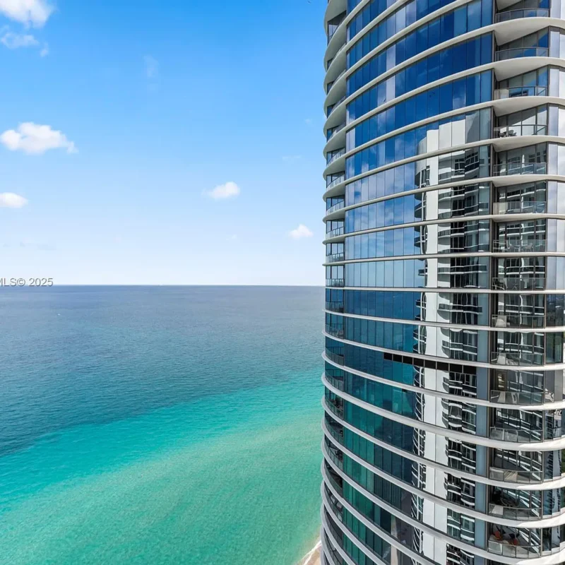 Curved glass high-rise condo tower along a turquoise sea and blue sky.
