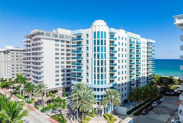 White modern condo building facing the ocean, with balconies and palm trees along a street. A blue sky and sea visible in the background.