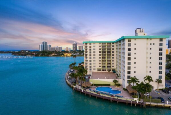 White apartment building by a marina with a curved dock, blue water, and a circular pool area. Palm trees line the foreground.