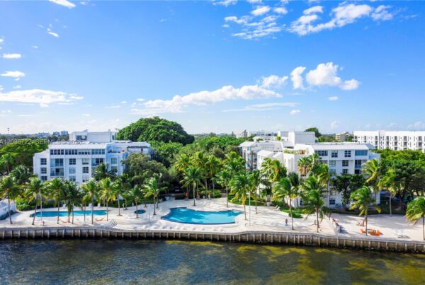 Aerial view of a white residential complex along a waterway, with a large oceanfront pool area surrounded by palm trees and modern buildings under a blue sky.