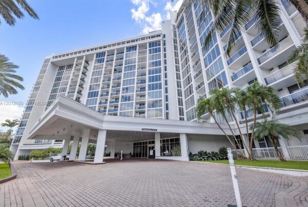 Wide view of a white, multi-story hotel/condo with glass balconies and a covered entrance, palm trees framing the scene under a blue sky