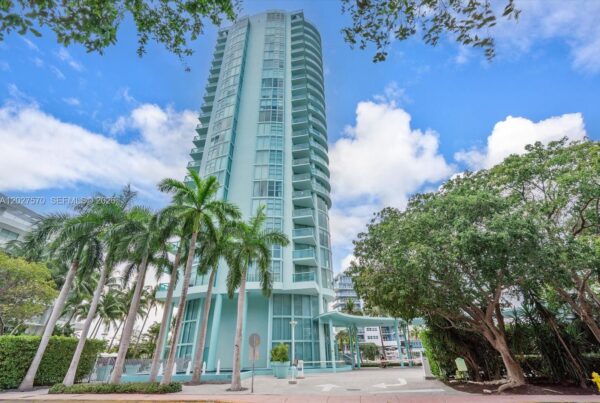 Turquoise high-rise condo with curved glass balconies, palm trees lining the sidewalk under a bright blue sky.